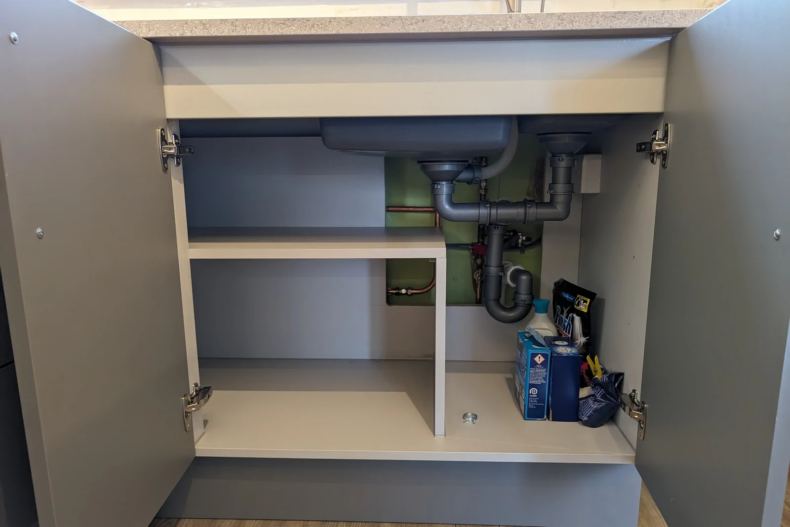 Inside view of the sink cabinet showing organised pipework, divider panel, and shelving for cleaning supplies of all sizes.