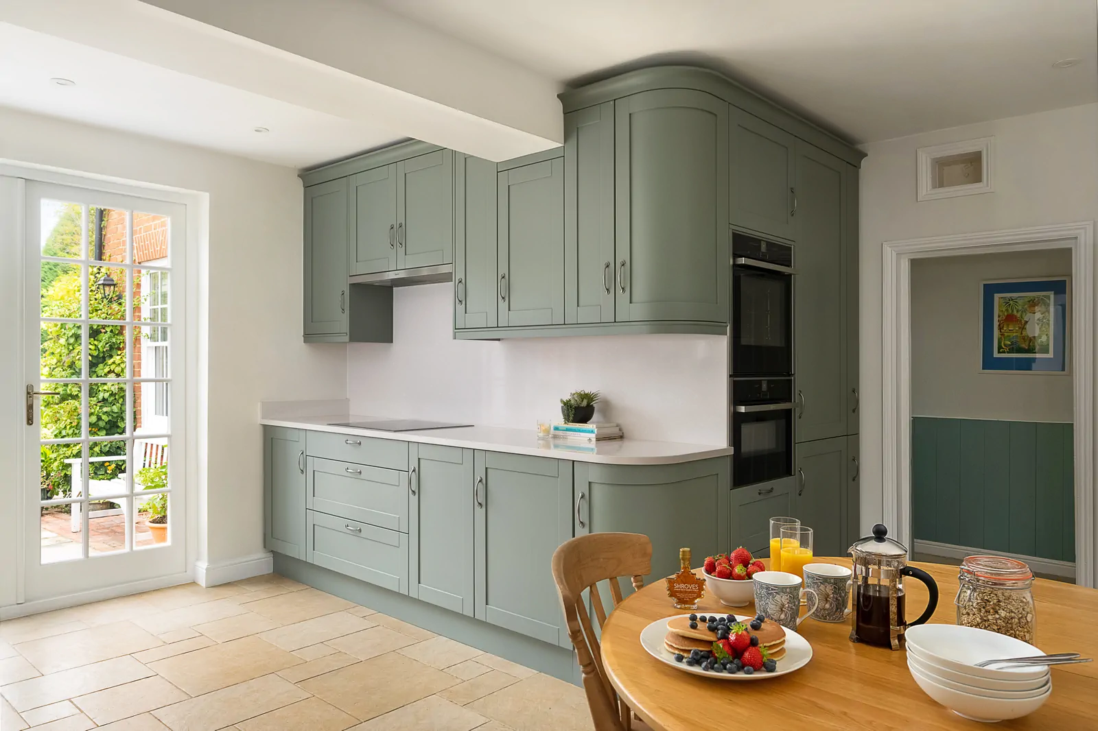 Wide view of a reed green shaker kitchen in Binfield with Neff ovens, Silestone worktops, and breakfast table, designed by Redwood Kitchens, Wokingham.