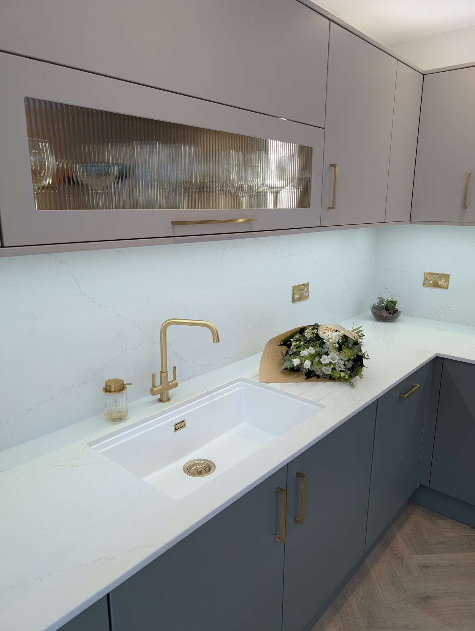 Blue-grey handleless kitchen with a white quartz worktop, brass tap and fluted glass wall cabinet